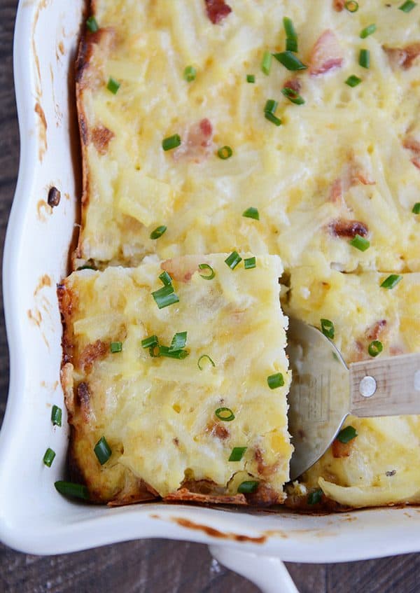 A dish full of cheesy egg casserole sprinkled with green onion. The front left piece is cut and being scooped out.