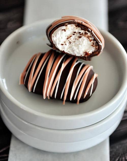 Two chocolate covered and chocolate drizzled marshmallow eggs on white plates. The top marshmallow egg has a bite taken out.