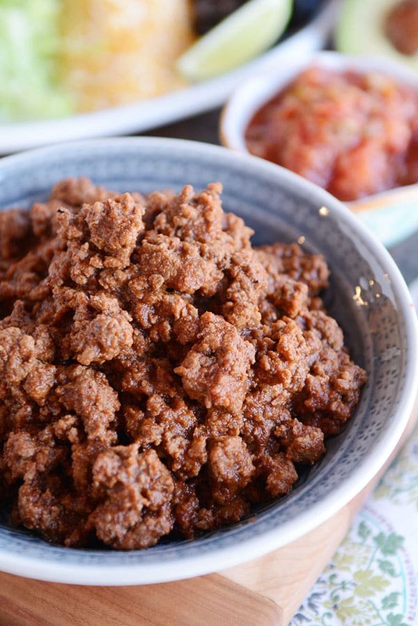 A bowl full of taco meat with a plate of toppings and a bowl of salsa in the background.