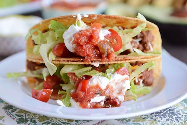 Two hard-shelled tacos stacked on top of each other, filled with ground beef, lettuce, tomatoes, and sour cream.